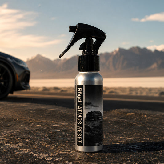 A silver spray bottle labeled Ryhd Atmos Reset  stands on a dark road with blurred mountains and a car in the background, suggesting an outdoor or automotive setting.